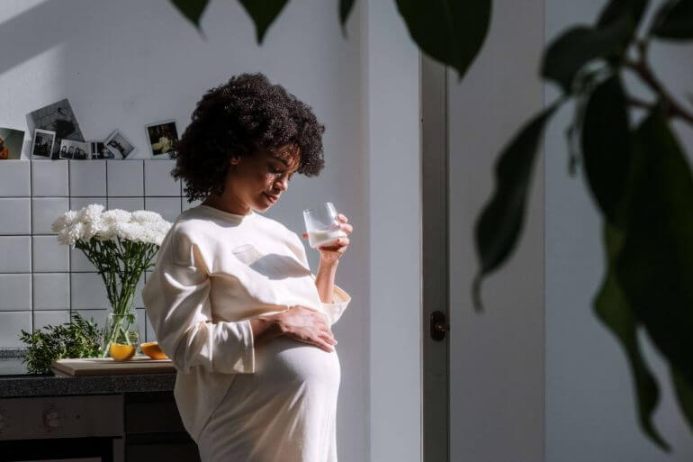 A pregnant individual drinking a glass of water with a hand on their stomach