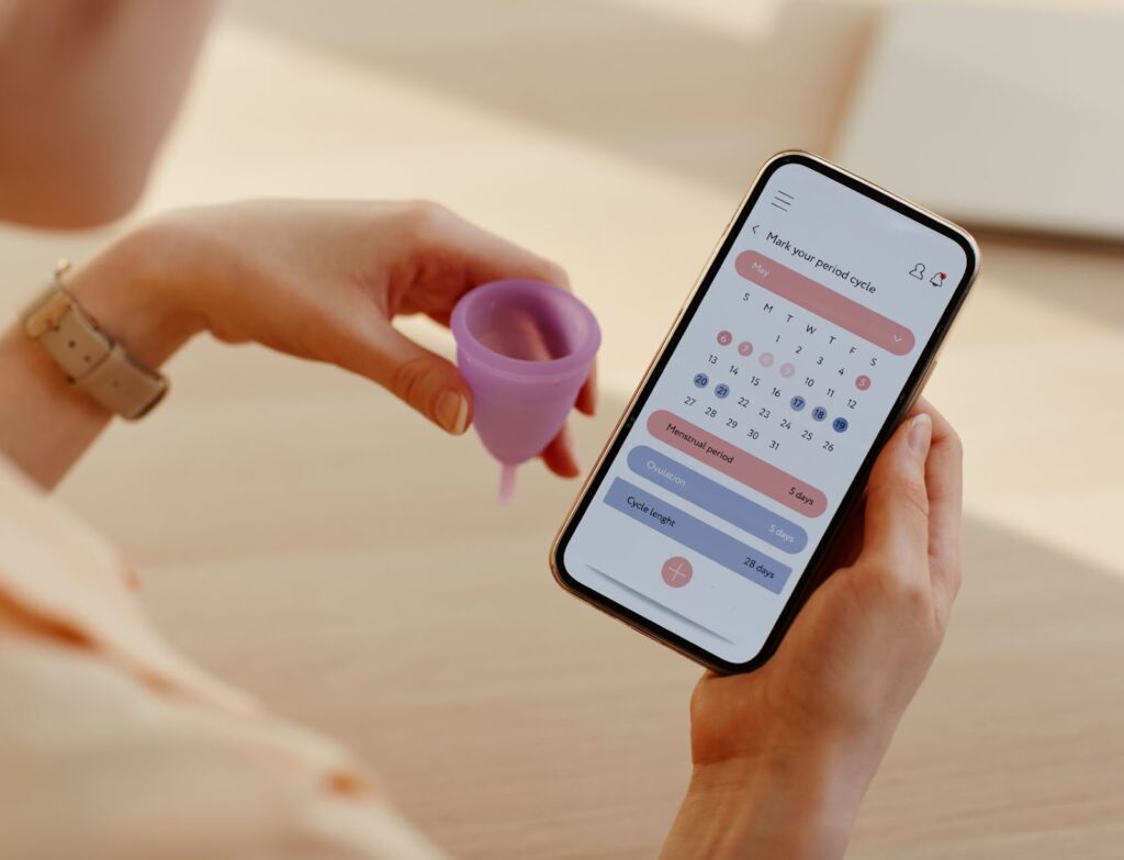 A woman holding a period cup while looking at a phone app to track their menstrual cycle.
