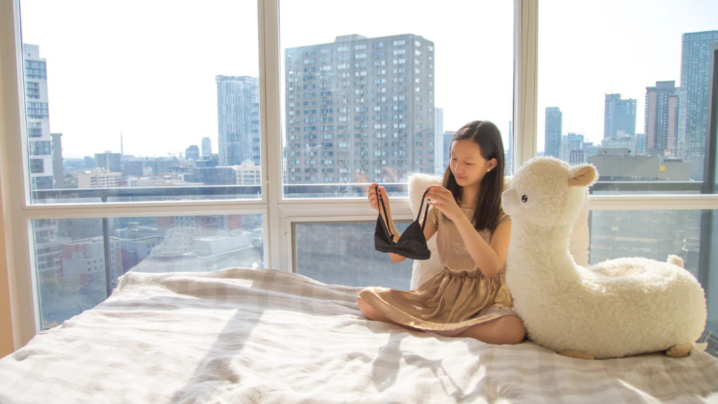 A young woman sitting on a bed in a sunlit room, holding a black training bra, with a large stuffed llama by her side. The room has large windows overlooking a cityscape.